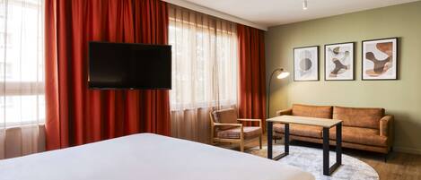 A hotel room with a bed, a TV, a sofa, a chair, and artwork on the wall.