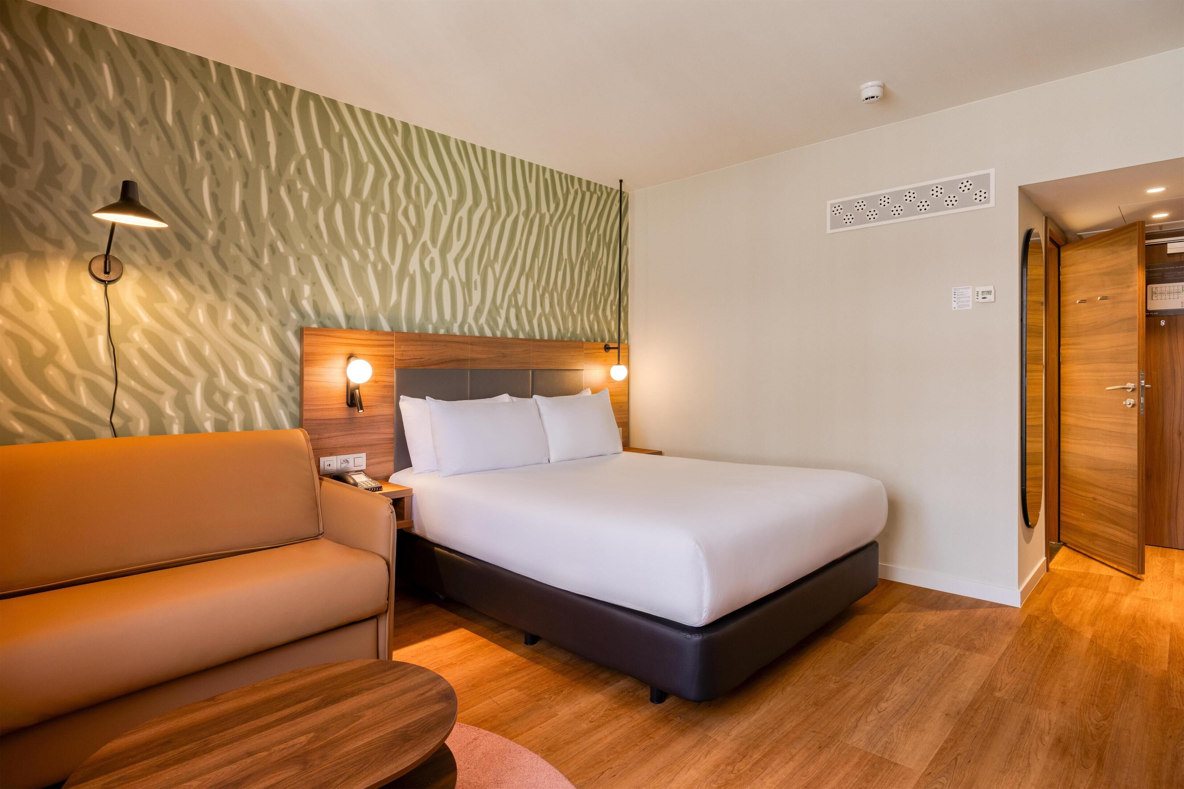 Habitación de hotel con cama, sofá, mesita de noche, lámpara y puerta.