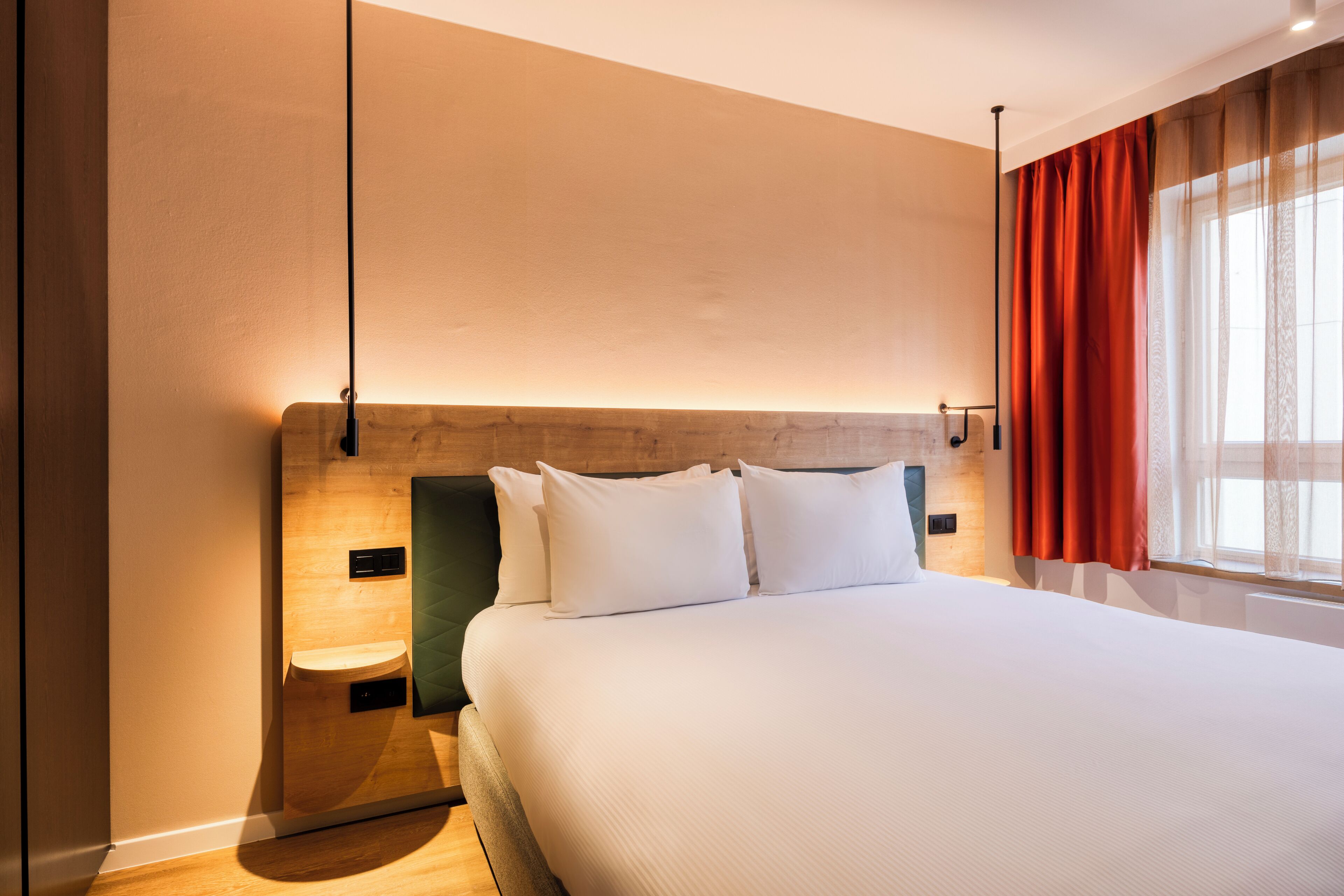 A hotel room with a large bed, a wooden headboard, a nightstand, and a window with curtains.