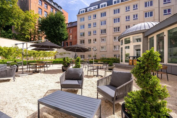 An outdoor seating area with wicker furniture and umbrellas, surrounded by a gravel surface and greenery.
