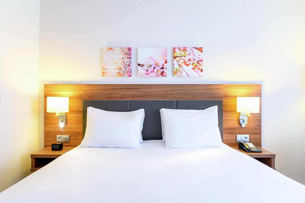 A hotel room with a large bed, two bedside tables with lamps, and three framed floral artworks on the wall.