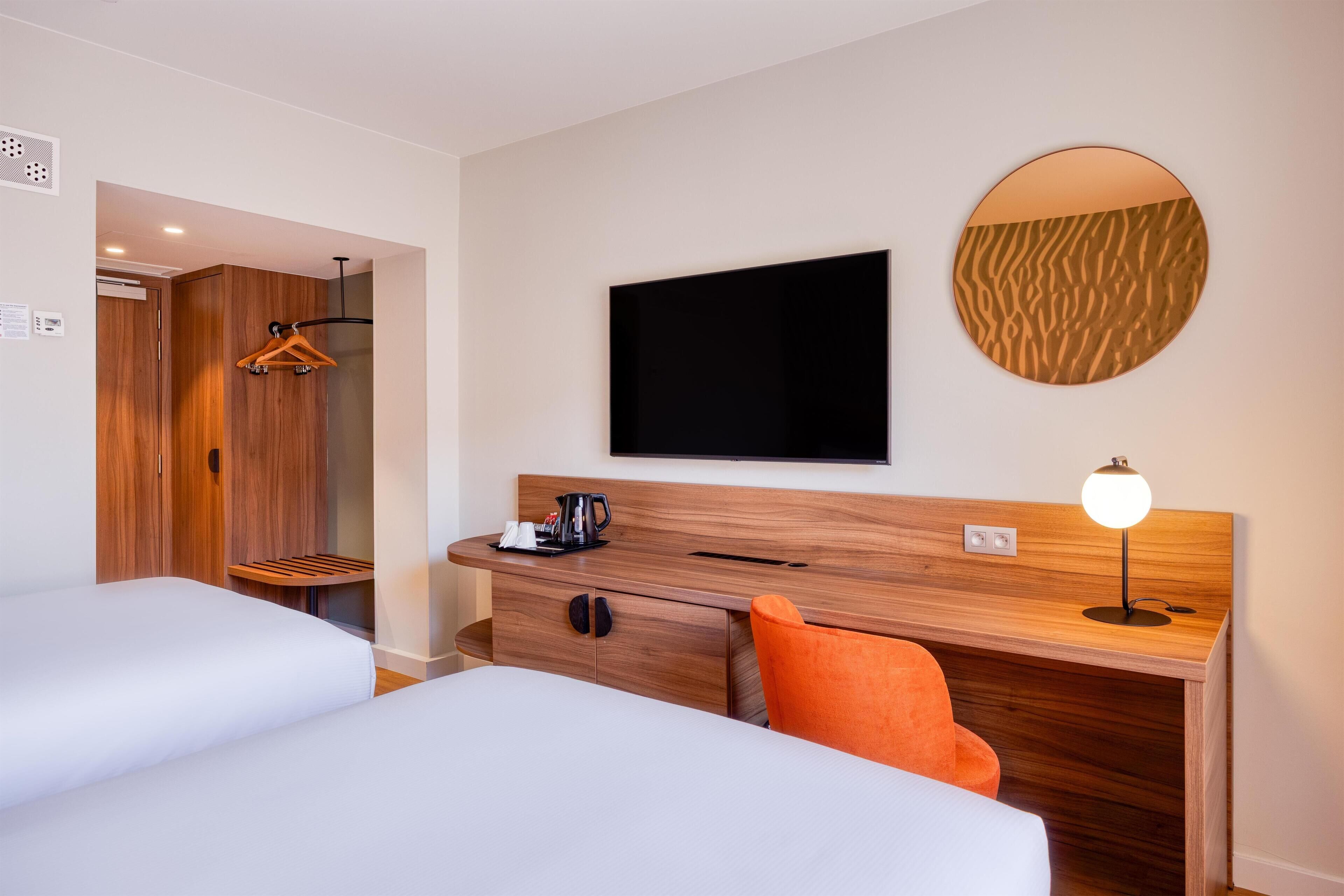 A hotel room with two beds, a desk with a lamp, a TV, and a mirror.