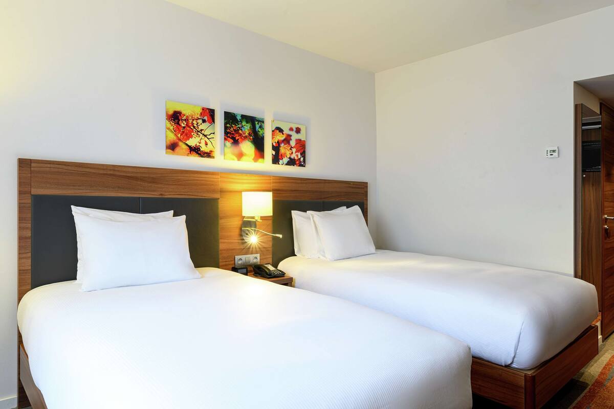 A hotel room with two beds, a wooden headboard, a nightstand with a lamp, and three wall-mounted artworks.