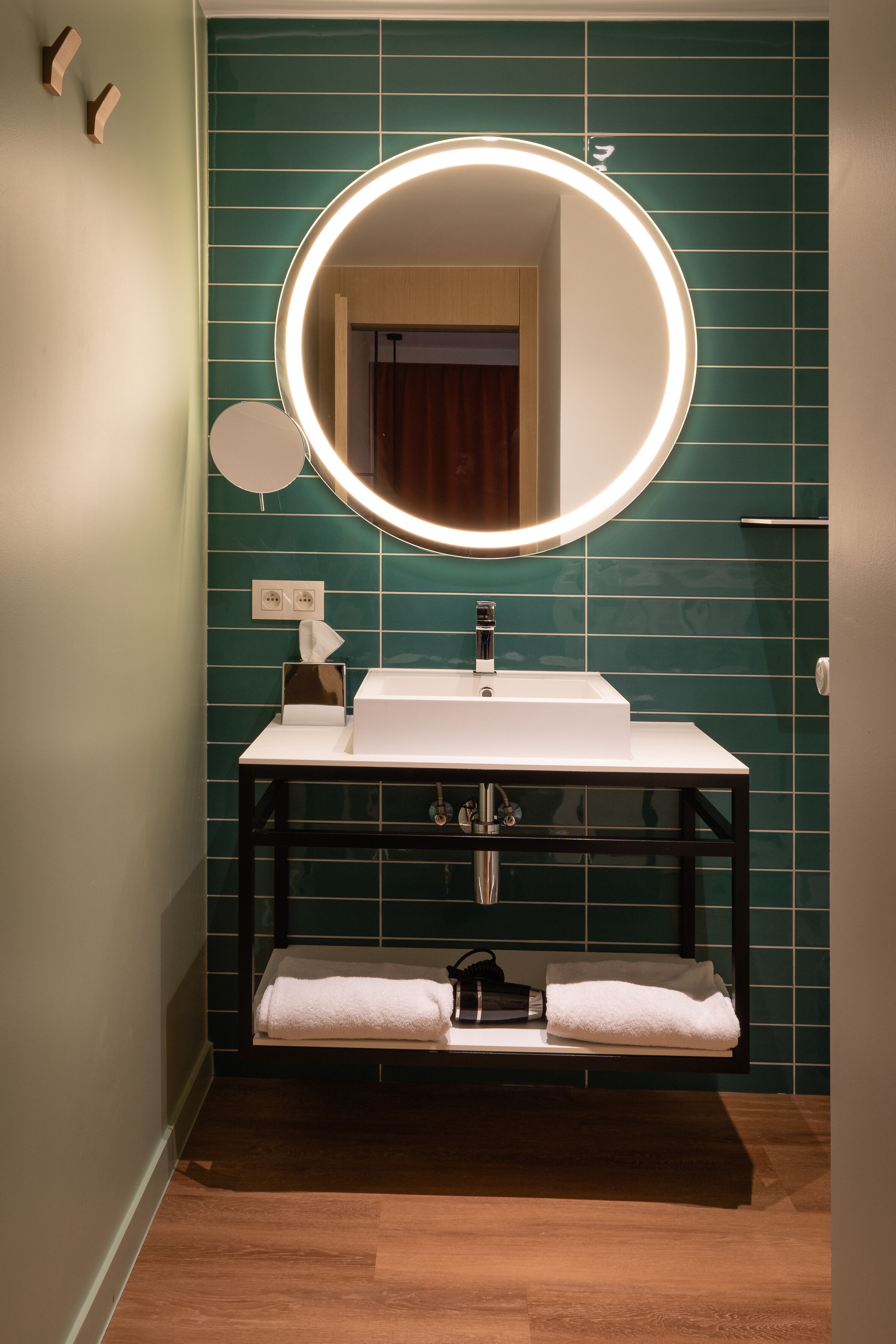 Un baño con espejo redondo, lavamanos blanco y paredes cubiertas de azulejos verdes.