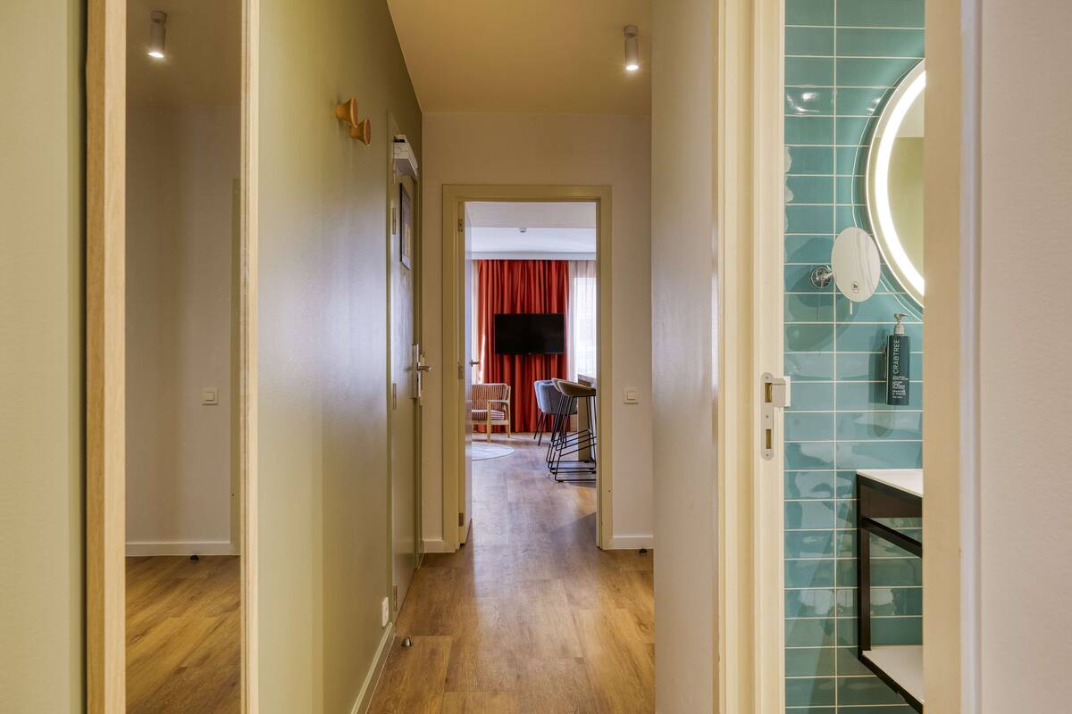 A hallway with wooden flooring leading to a room with a red curtain and a flat-screen TV.