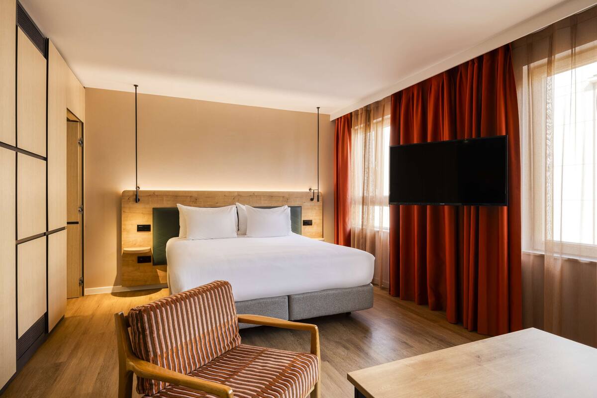 A hotel room with a large bed, a TV mounted on the wall, a wooden chair, and a wooden table.