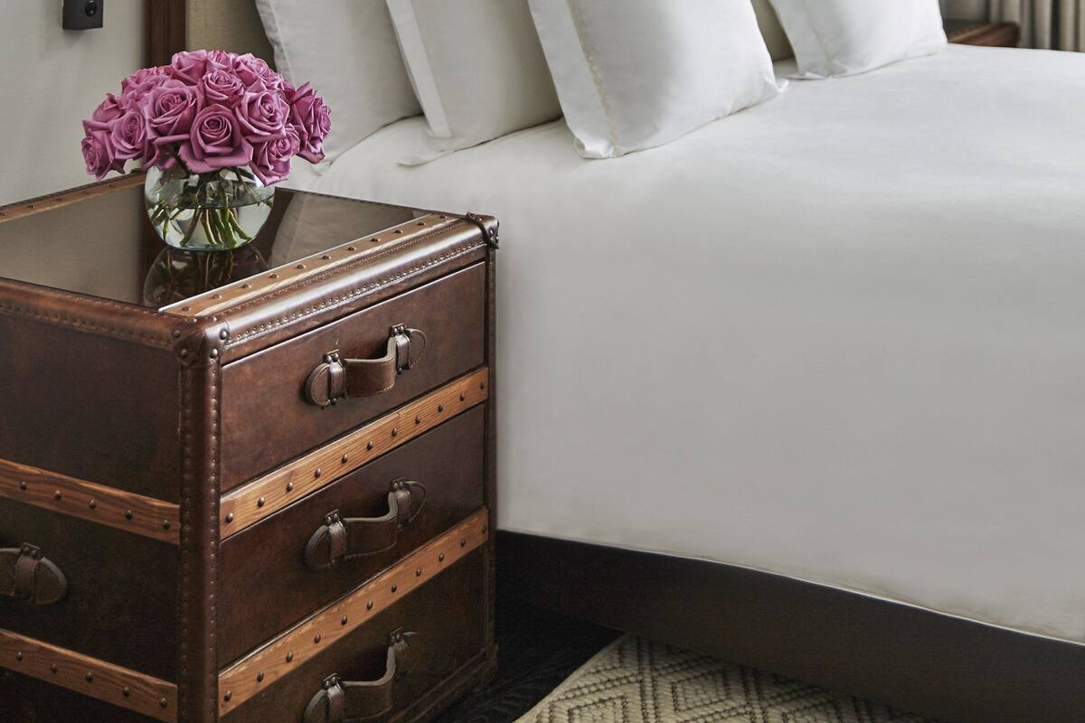 Un lit bien fait, avec du linge de lit blanc, une table de chevet en bois dotée de tiroirs et un vase rempli de fleurs violettes.