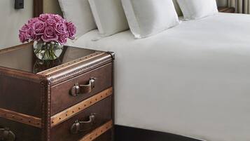 A neatly made bed with white linens, a wooden nightstand with drawers, and a vase of purple flowers.