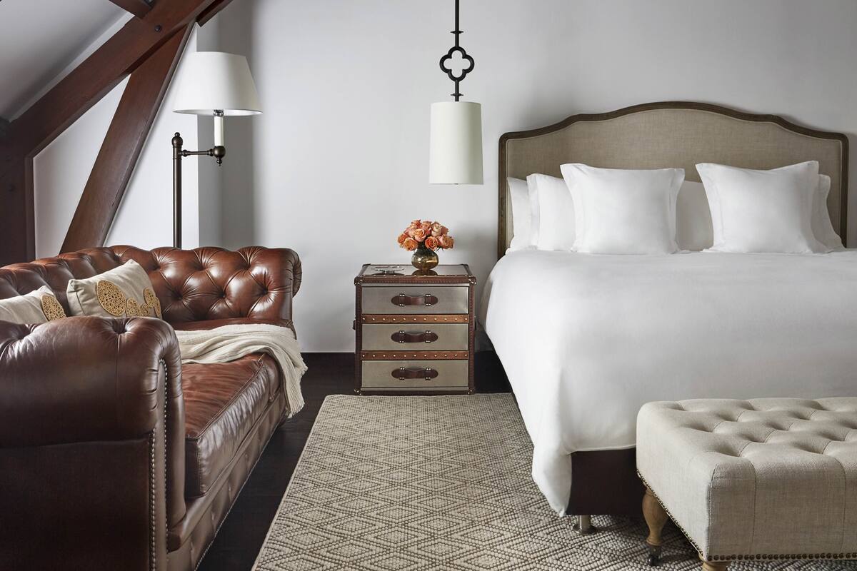 Une chambre avec un canapé en cuir marron, une table de chevet en bois avec une lampe, un lit avec du linge de lit blanc et un tapis à motifs.