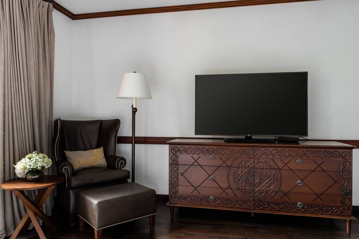 Une chambre d’hôtel avec un fauteuil en cuir, une table d’appoint en bois avec une lampe, un téléviseur à écran plat et un meuble décoratif en bois.