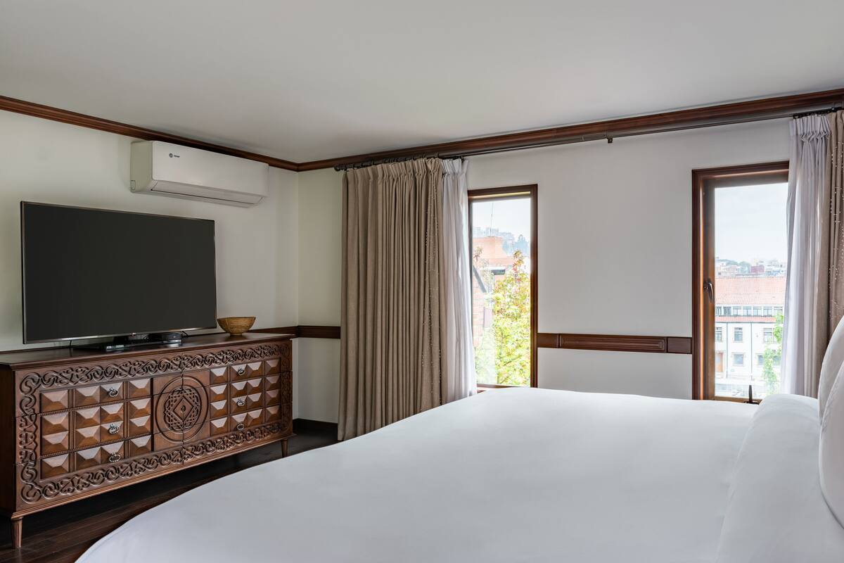 Une chambre d’hôtel avec un grand lit, un téléviseur posé sur une commode en bois, un balcon avec des rideaux et une vue sur la ville.