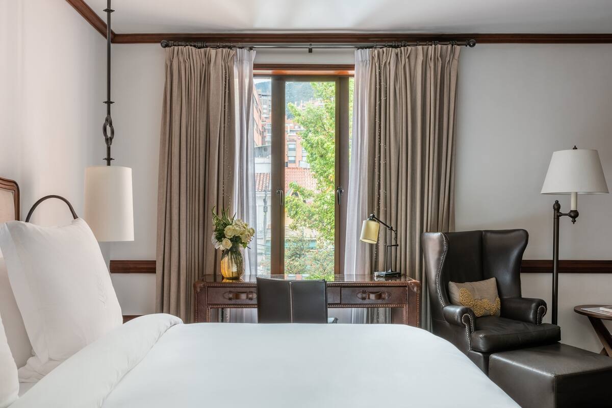 Une chambre d’hôtel avec un lit, un bureau, une chaise, une lampe et une vue sur les bâtiments par la fenêtre.