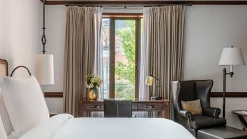 A hotel room with a bed, a desk, a chair, a lamp, and a view of buildings through the window.