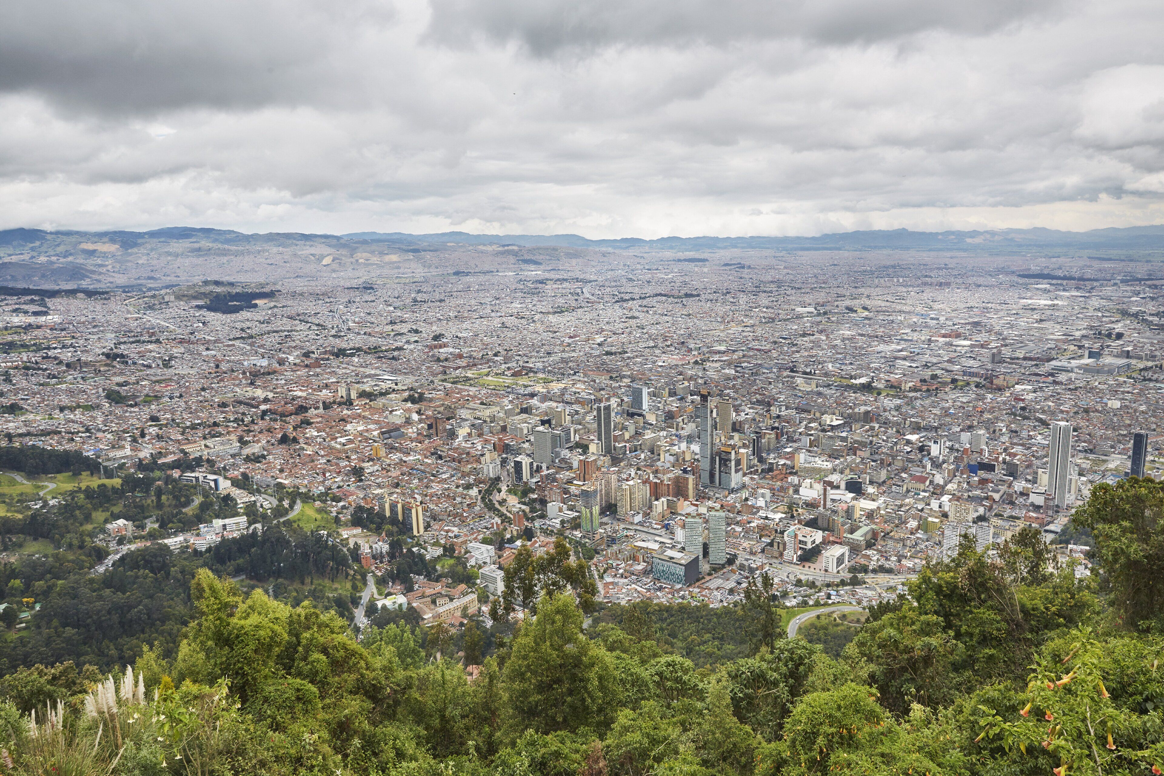 Une vue panoramique d’une ville densément peuplée, avec un mélange d’immeubles de grande hauteur et de bâtiments plus petits, entourée de verdure et sous un ciel nuageux.