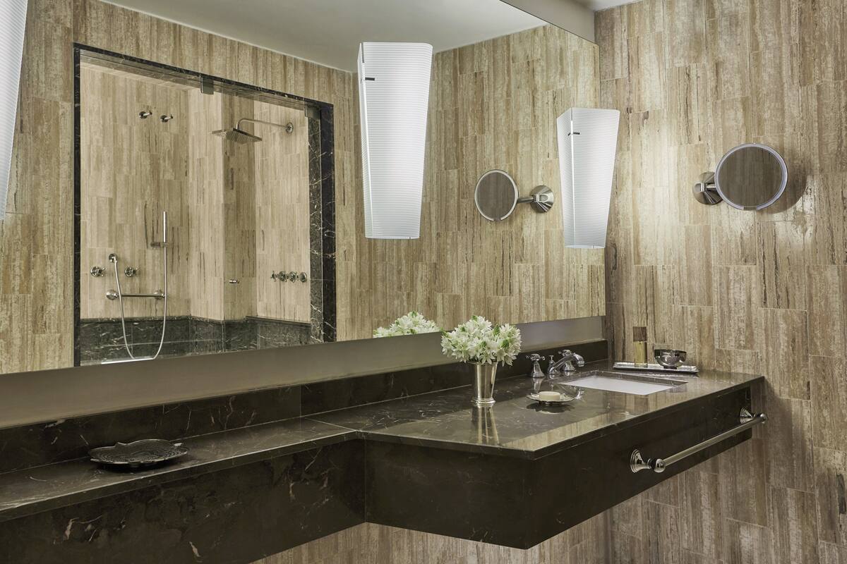 Une salle de bain moderne avec un grand miroir, un plan de toilette noir, un espace douche et un vase rempli de fleurs.