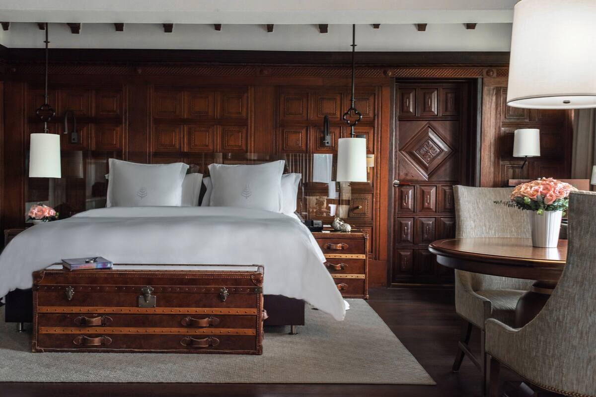 Une chambre à coucher avec un grand lit, des lambris en bois, une vieille malle et une table sur laquelle est posé un vase.