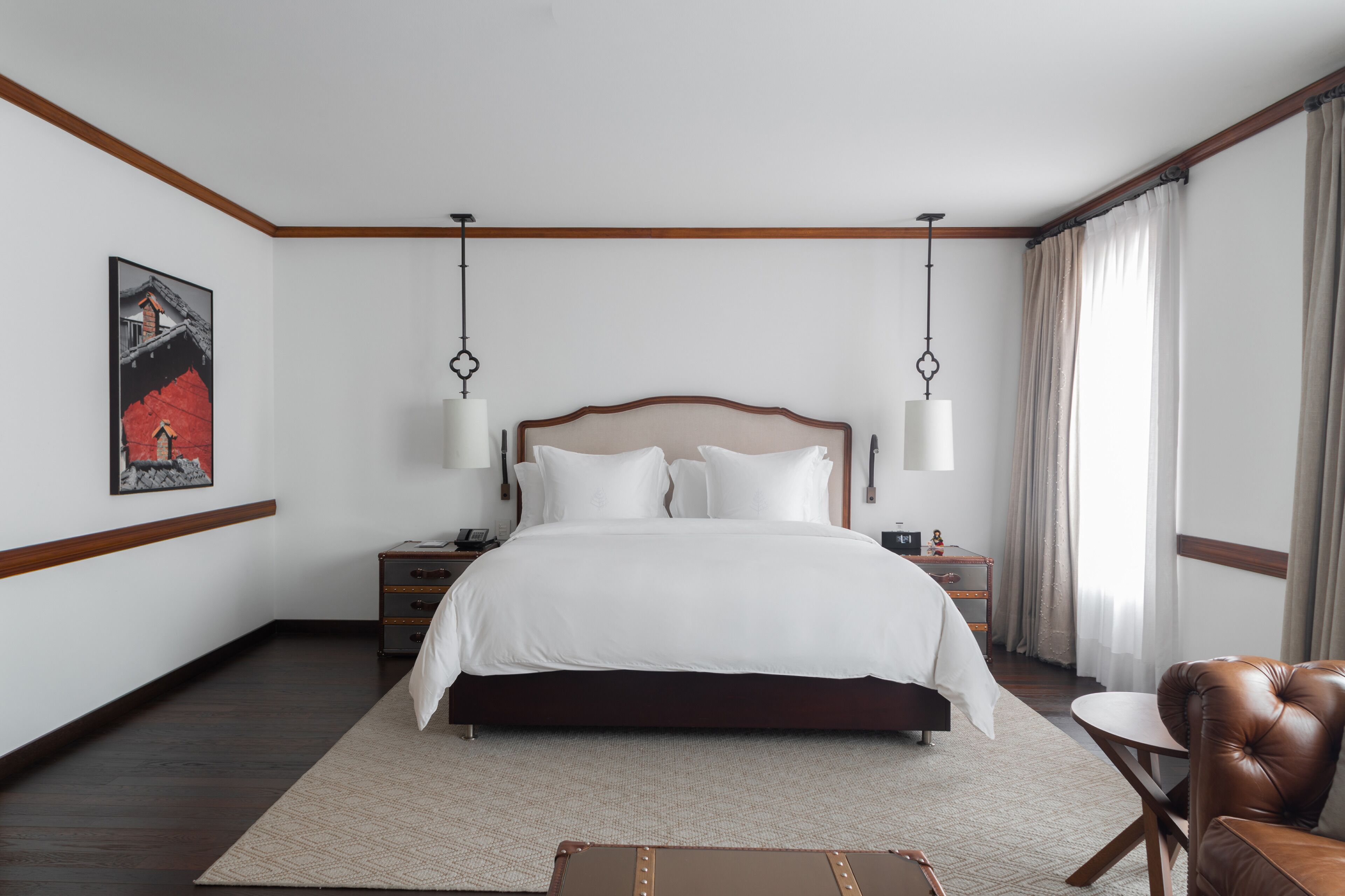 A hotel room with a large bed, two bedside tables, a brown leather armchair, a small round table, and a painting on the wall.