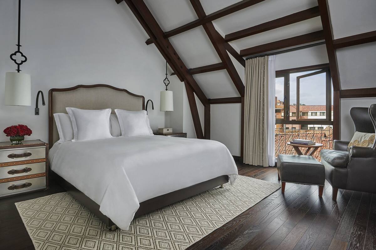 Une chambre avec un grand lit, une table de chevet, une chaise, une fenêtre donnant sur des immeubles et un plafond en bois.