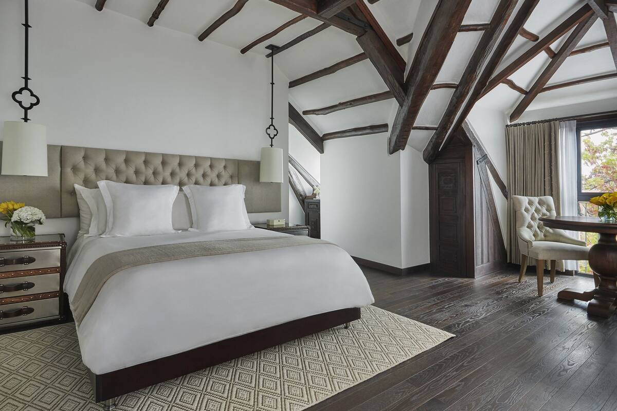 Une chambre avec un grand lit, une table de chevet, une commode, une fenêtre avec des rideaux et un coin repas comprenant une table et des chaises.