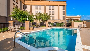Una piscina rectangular con una escalera metálica, rodeada de sillas de descanso y un edificio con varias ventanas al fondo.