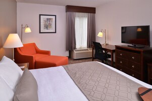 Una habitación de hotel con cama, escritorio, silla, televisor y ventana con cortinas.