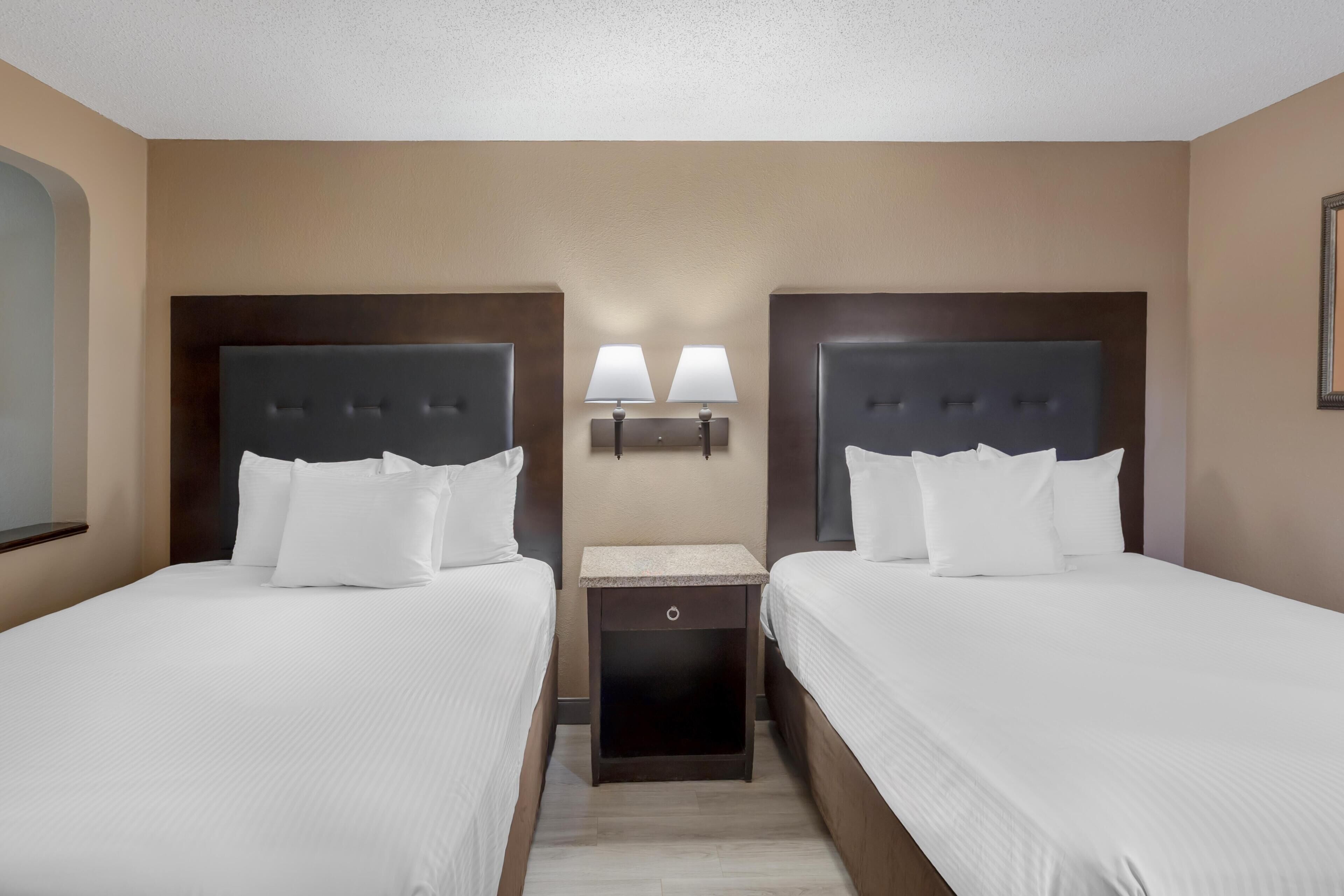 A hotel room with two beds, a nightstand, and two wall-mounted lamps.