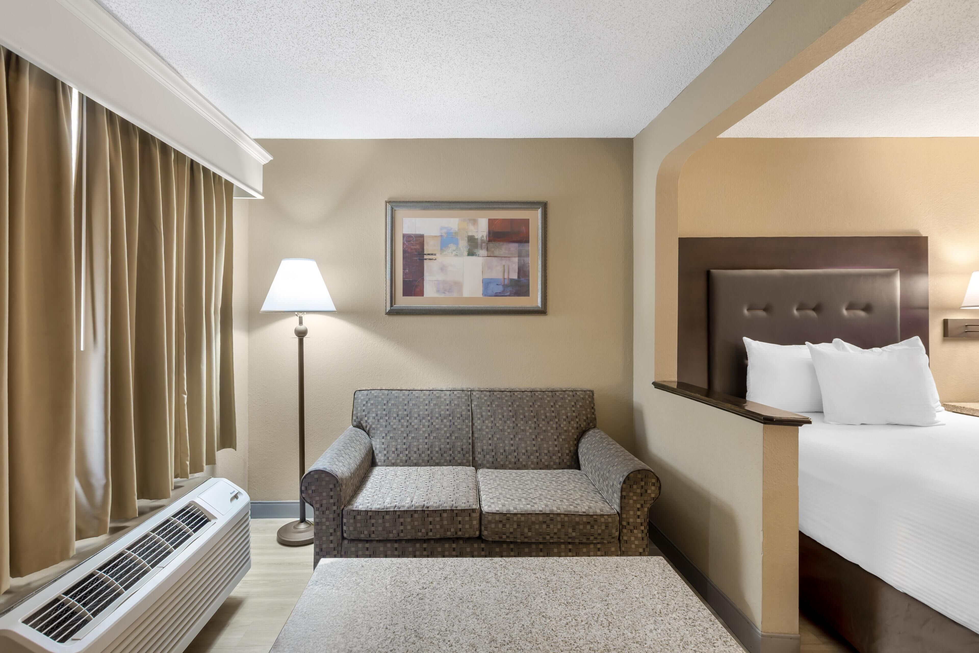 A hotel room with a bed, a sofa, a standing lamp, a framed artwork, and a window with curtains.