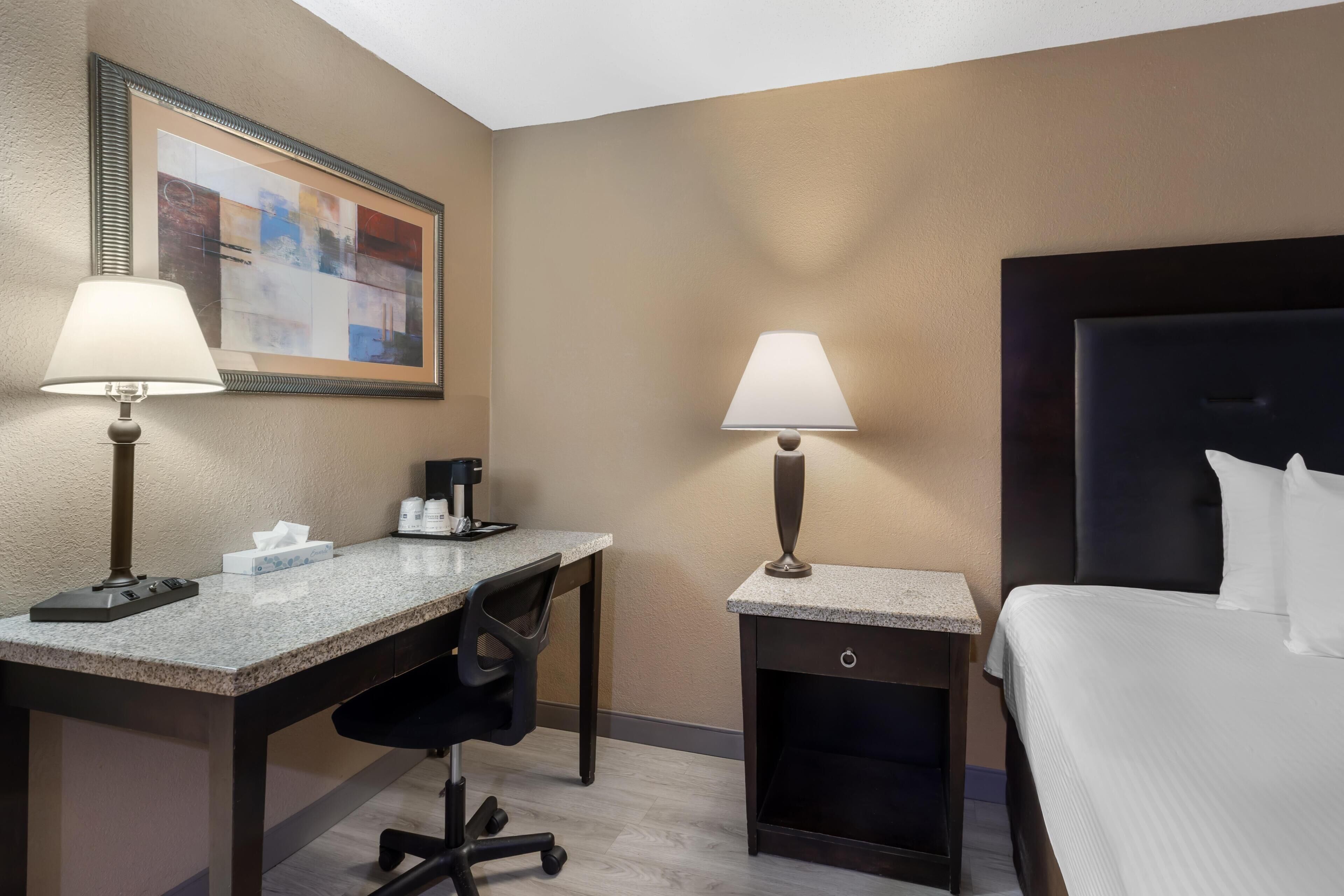 A hotel room with a desk, two lamps, a chair, a bed, and a framed artwork on the wall.
