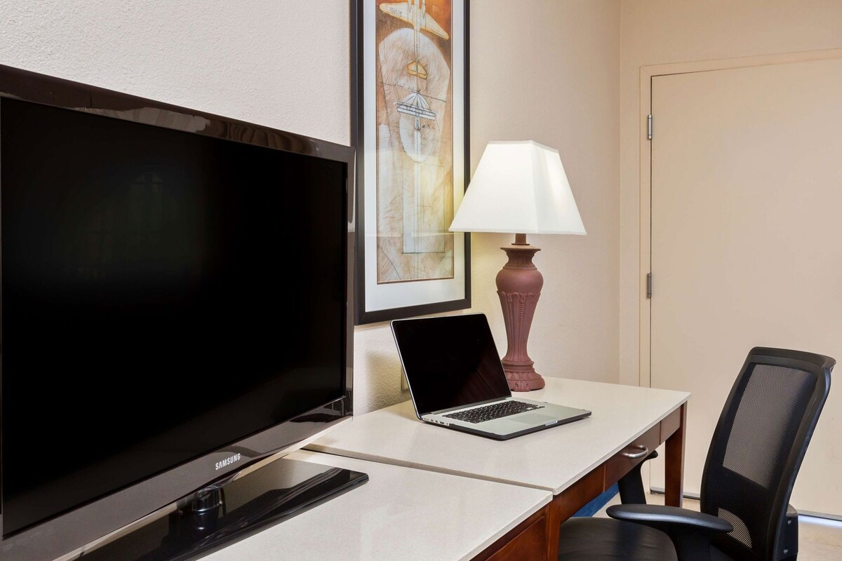 Ein Hotelzimmer mit Flachbildfernseher, einem Laptop auf einem Schreibtisch, einer Lampe und einem Sessel.