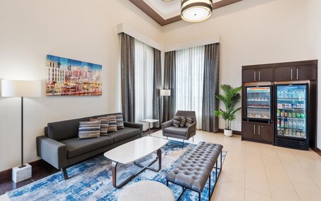 A modern living room with a sofa, armchair, coffee table, and ottoman. There is a large painting on the wall, a floor lamp, and a refrigerator with a Pepsi logo.