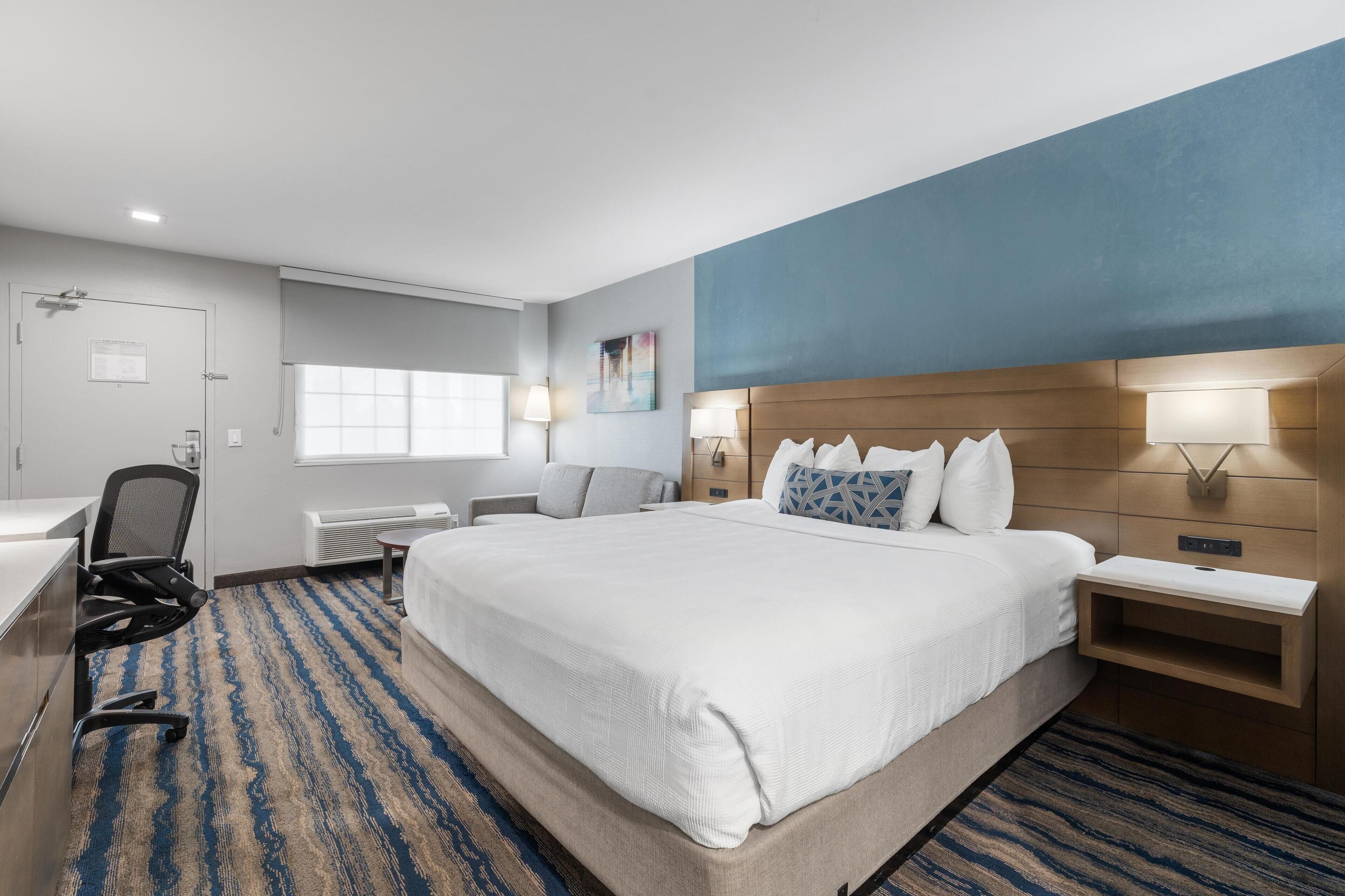 A hotel room with a large bed, a desk, a chair, a nightstand, and a wall-mounted lamp.