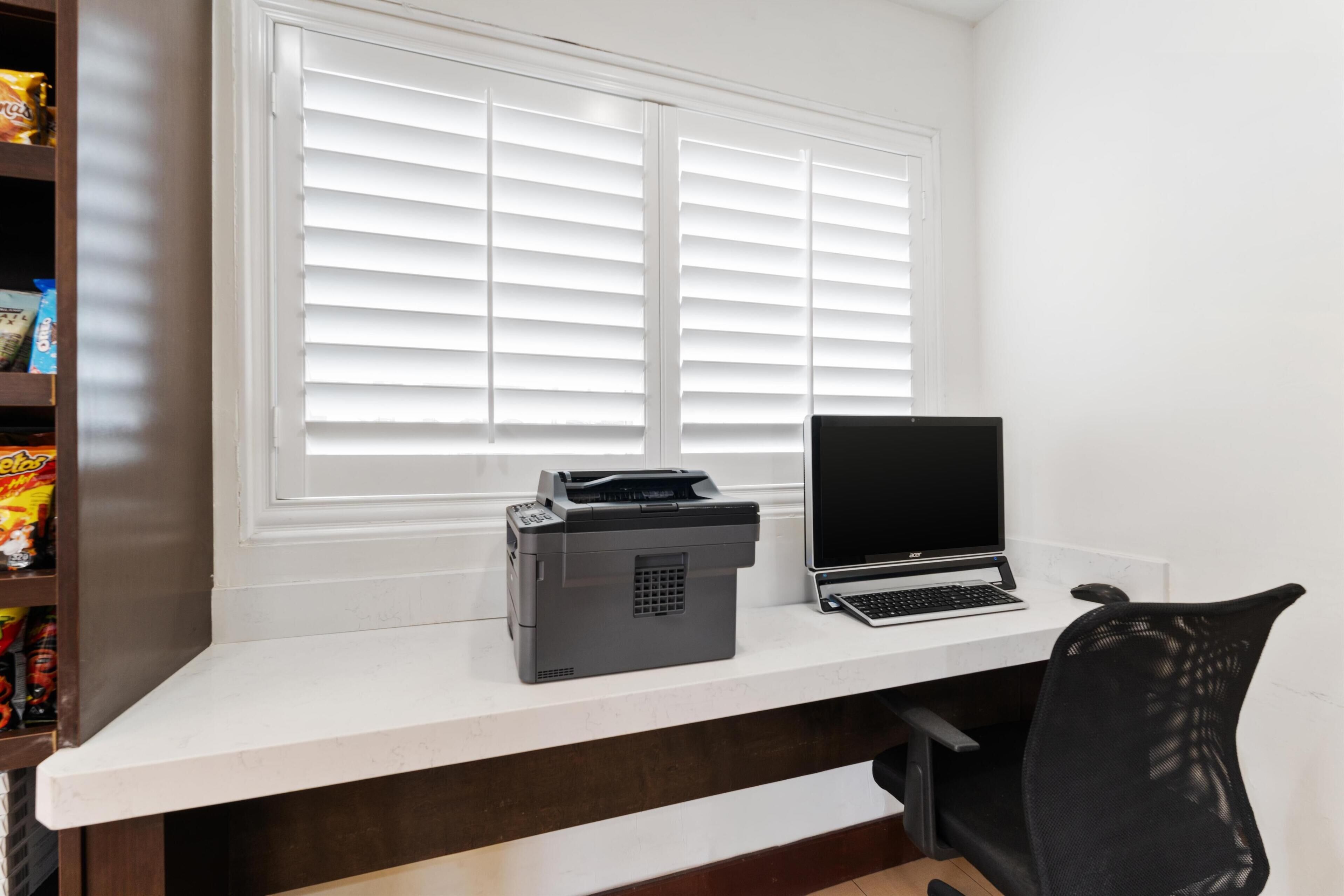 A workspace with a printer and laptop on a desk, a window with blinds, and a shelf with snacks.
