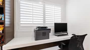 A workspace with a printer and laptop on a desk, a window with blinds, and a shelf with snacks.