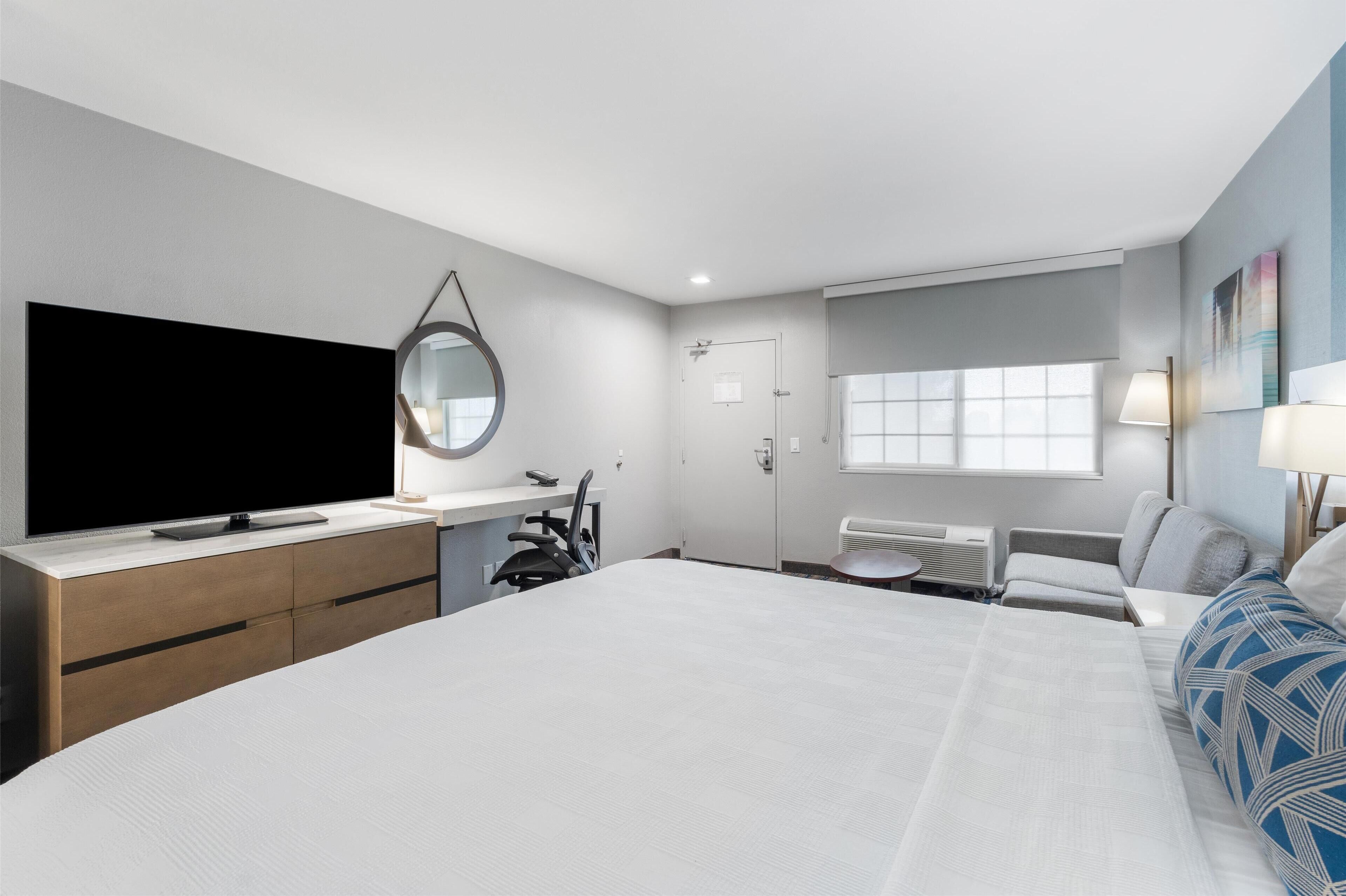 Une chambre d’hôtel moderne dotée d’un grand lit, d’un téléviseur à écran plat, d’un bureau avec une chaise, d’une petite table, d’une chaise, d’une fenêtre avec des stores et d’une lampe fixée au mur.