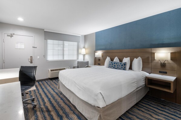 A hotel room with a large bed, a desk, a chair, a nightstand, and a window with blinds.