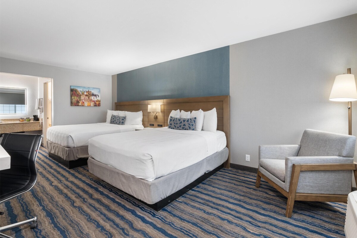Une chambre d’hôtel avec deux lits, un bureau, une chaise, une lampe et un tableau au mur.