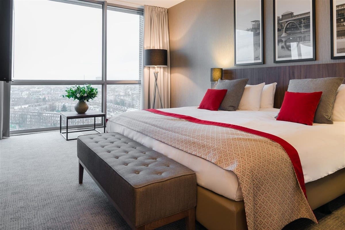 A modern hotel room with a large bed, a bench, a nightstand, and a view of the city through large windows.