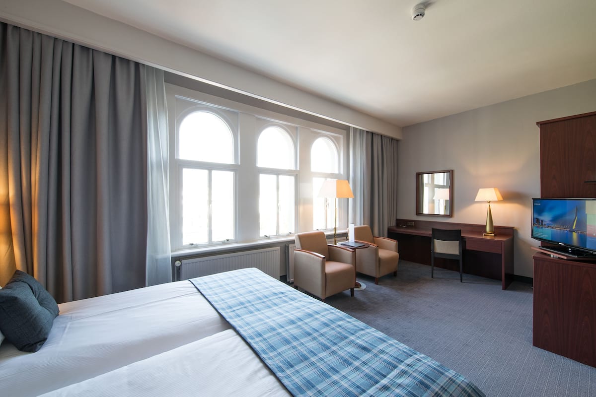 A hotel room with a large bed, two armchairs, a desk, a television, and a window with curtains.