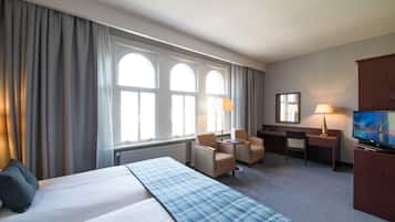 Een hotelkamer met een groot bed, twee fauteuils, een bureau, een televisie en een raam met gordijnen.