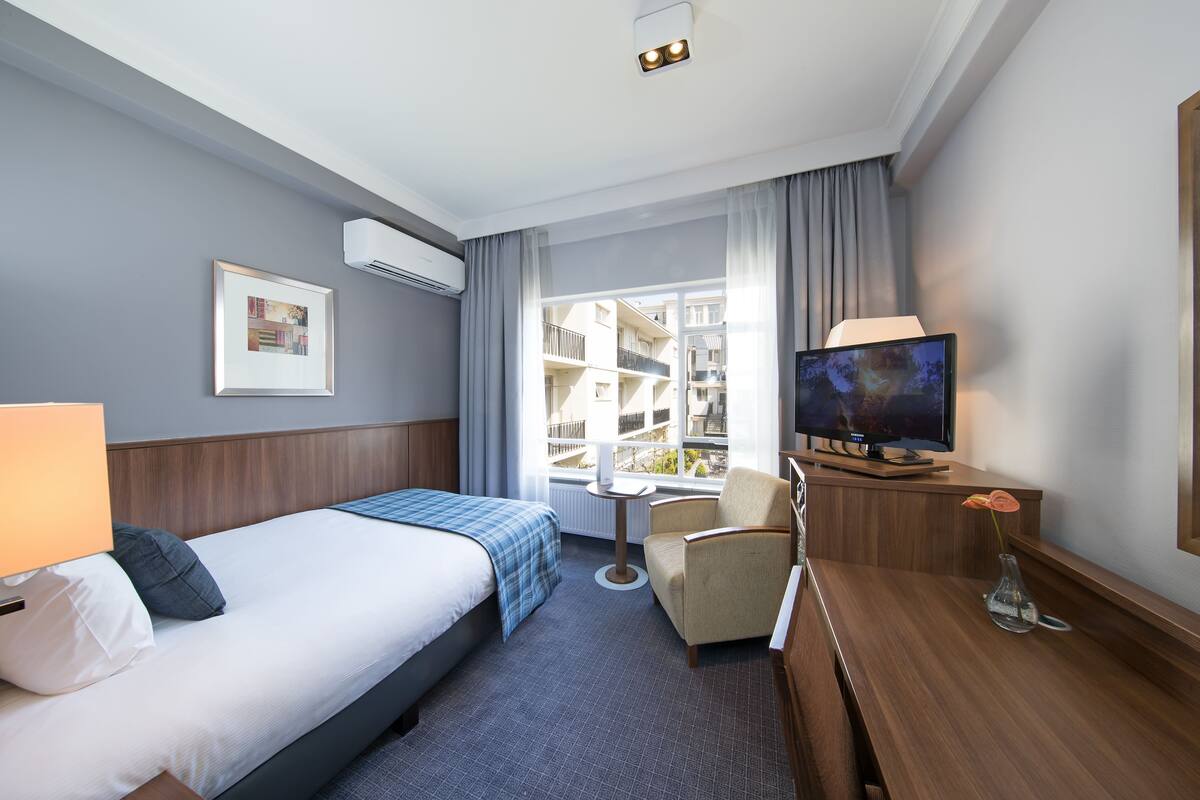 A hotel room with a bed, a desk with a TV, a chair, a small table, and a window with a view of buildings.