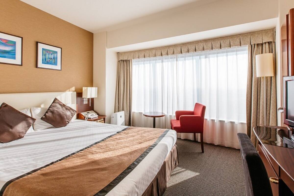Ein Hotelzimmer mit einem großen Bett, einem Schreibtisch mit Stuhl, einem Fernseher und einem Fenster mit Vorhängen.