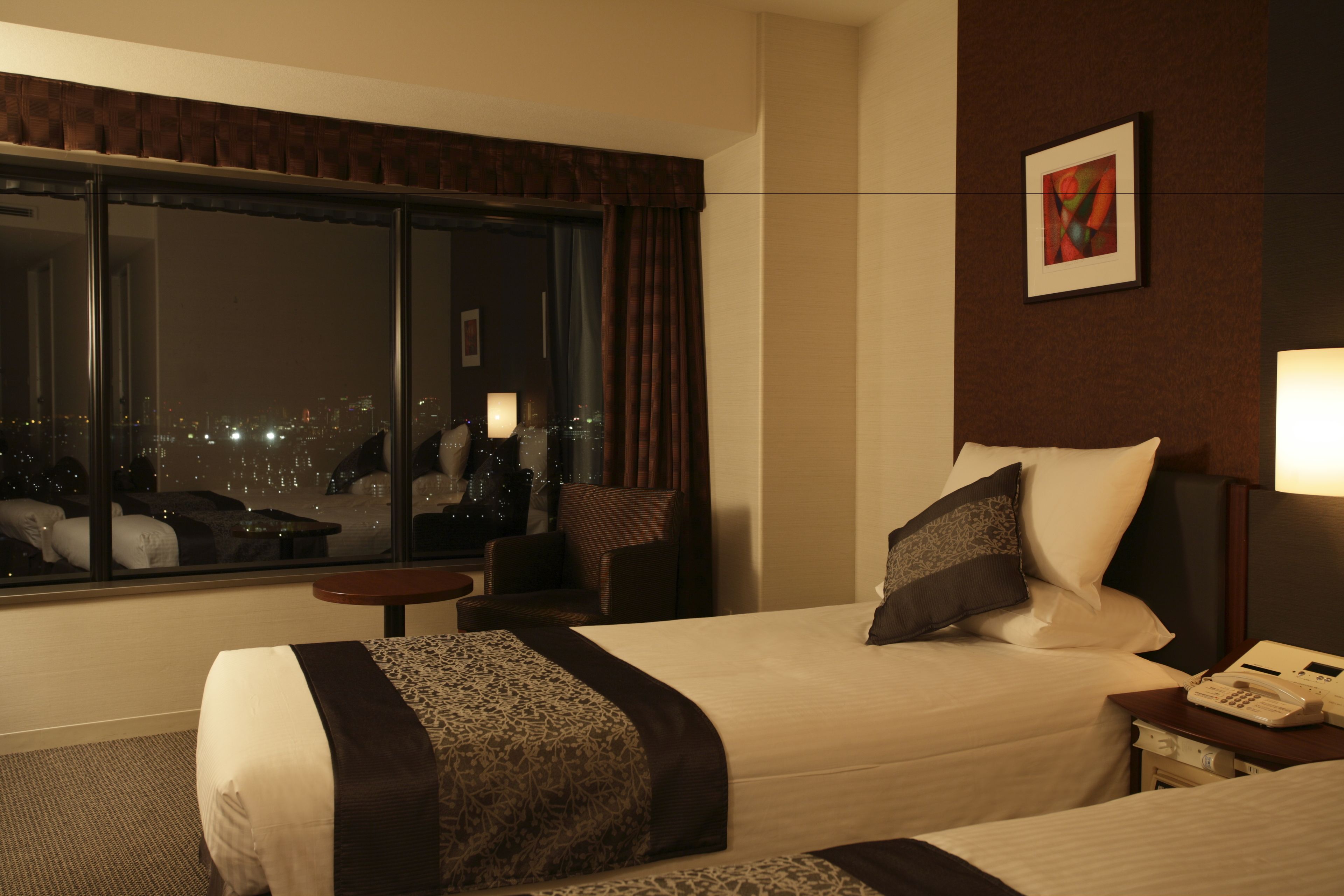 A hotel room with a large window, a bed with a patterned bedspread, a bedside table with a lamp, and a painting on the wall.