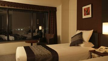 A hotel room with a large window, a bed with a patterned bedspread, a bedside table with a lamp, and a painting on the wall.