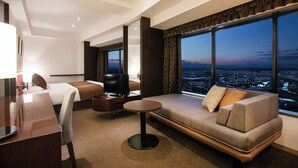 Une chambre d’hôtel moderne dotée d’une grande fenêtre donnant sur la ville, d’un canapé, d’un lit, d’une télévision et d’un bureau avec une lampe.