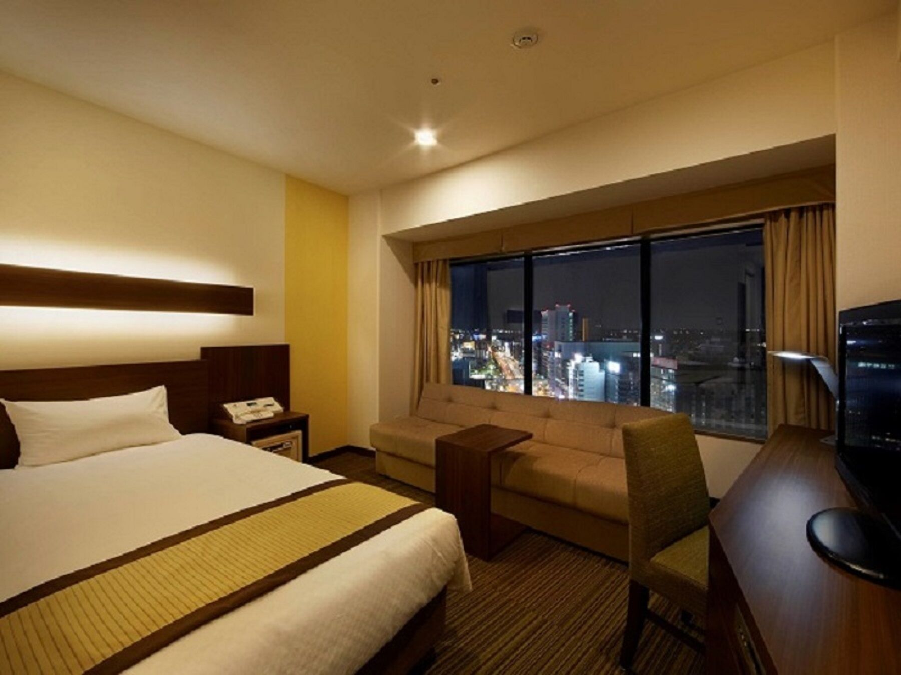 A hotel room with a bed, a sofa, a desk, and a large window offering a city view.