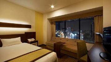 A hotel room with a bed, a sofa, a desk, and a large window offering a city view.