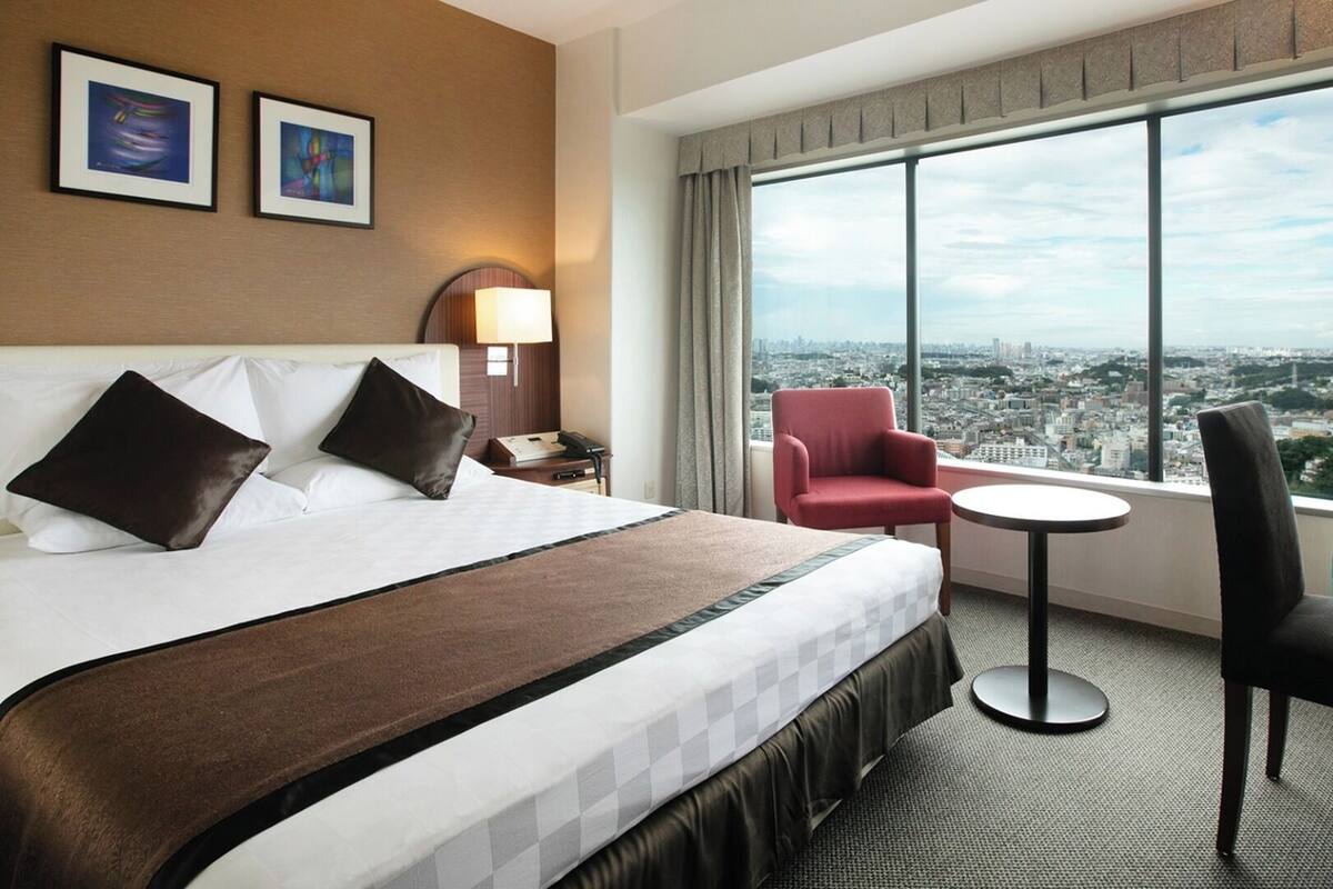 Ein Hotelzimmer mit einem großen Bett, einem roten Sessel, einem kleinen runden Tisch und Blick auf die Stadt durch große Fenster.