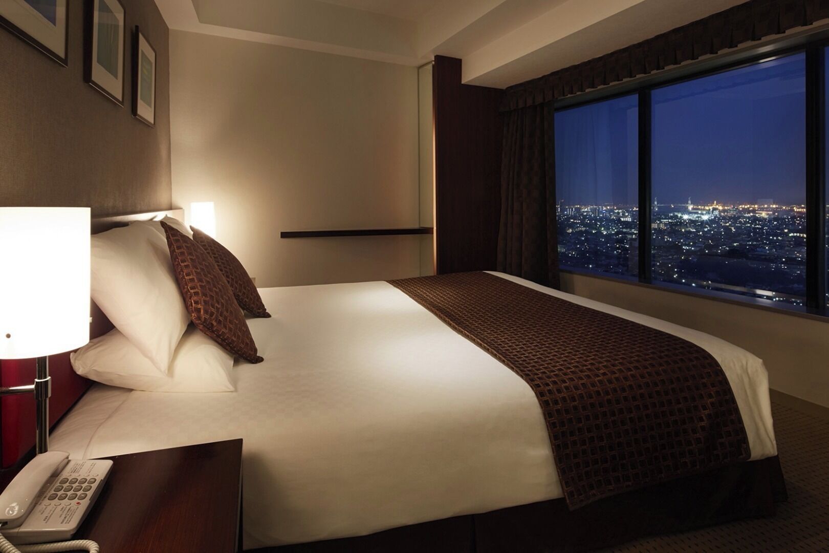 A hotel room with a large bed, a bedside telephone, a window with a city view, and framed pictures on the wall.