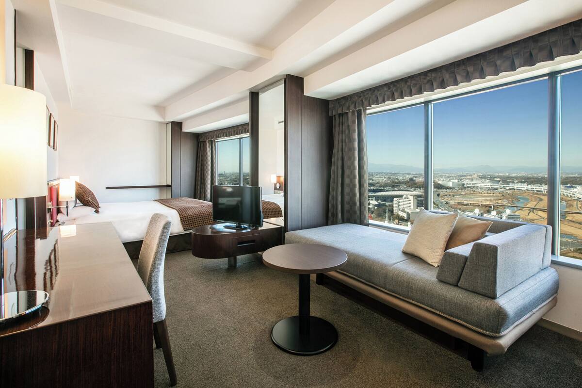 Ein Hotelzimmer mit Bett, Sofa, Fernseher, Schreibtisch und Blick auf die Stadt.