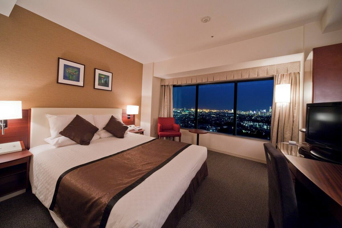 Ein Hotelzimmer mit einem großen Bett, einem roten Sessel, einem Schreibtisch mit Fernseher und Blick auf eine nächtliche Skyline.